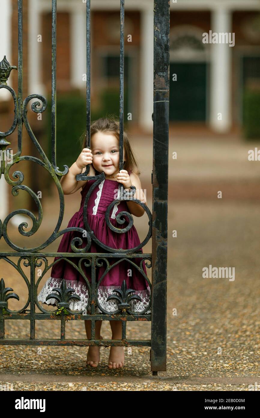 Beautiful two year old girl with a purple dress outside near an iron gate peeking through the gate Stock Photo