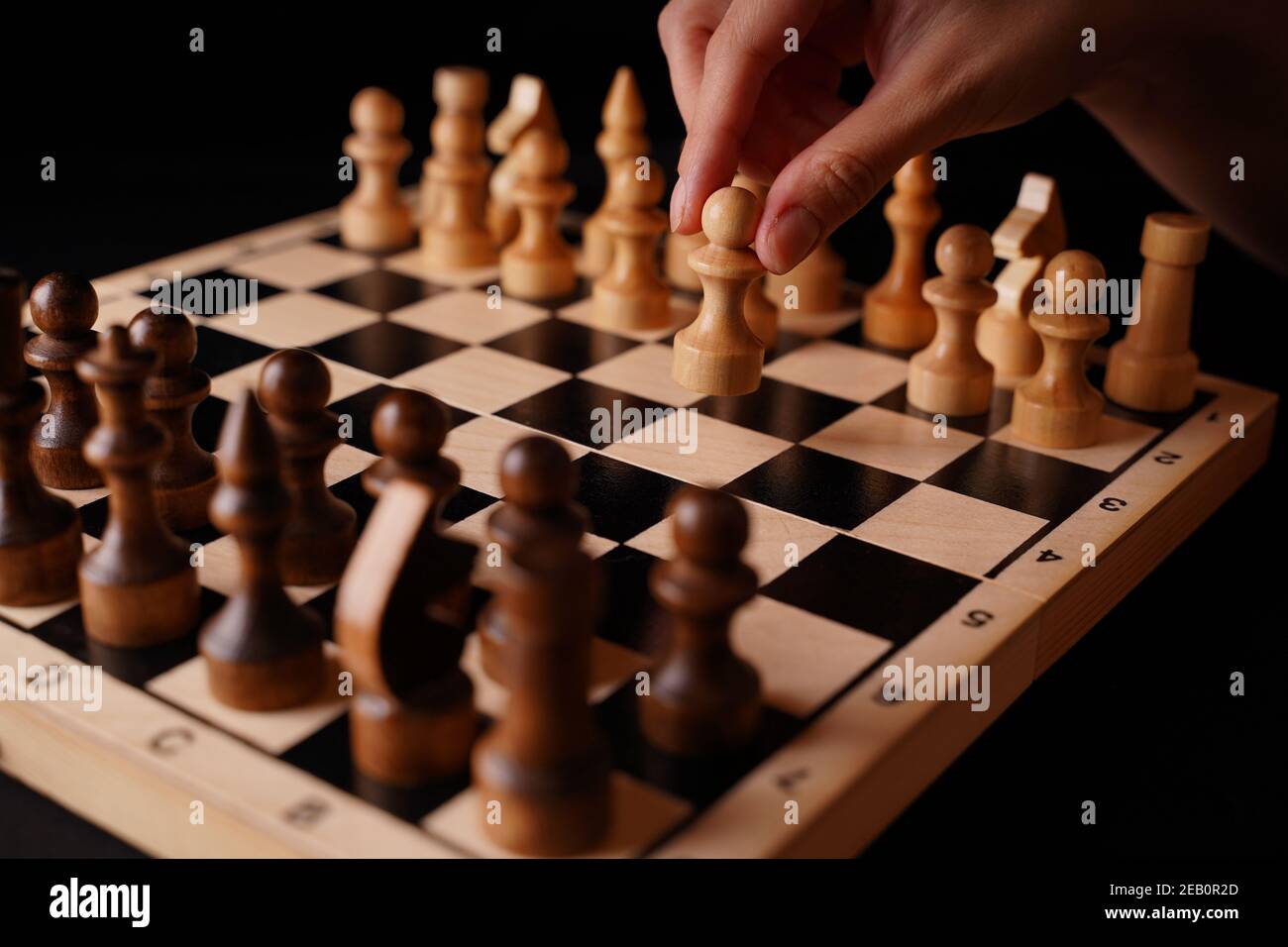 Close up of white and black wooden chess pieces on board. Woman's hand makes first move of white pawn on chessboard. Concept of intelligent, logical a Stock Photo