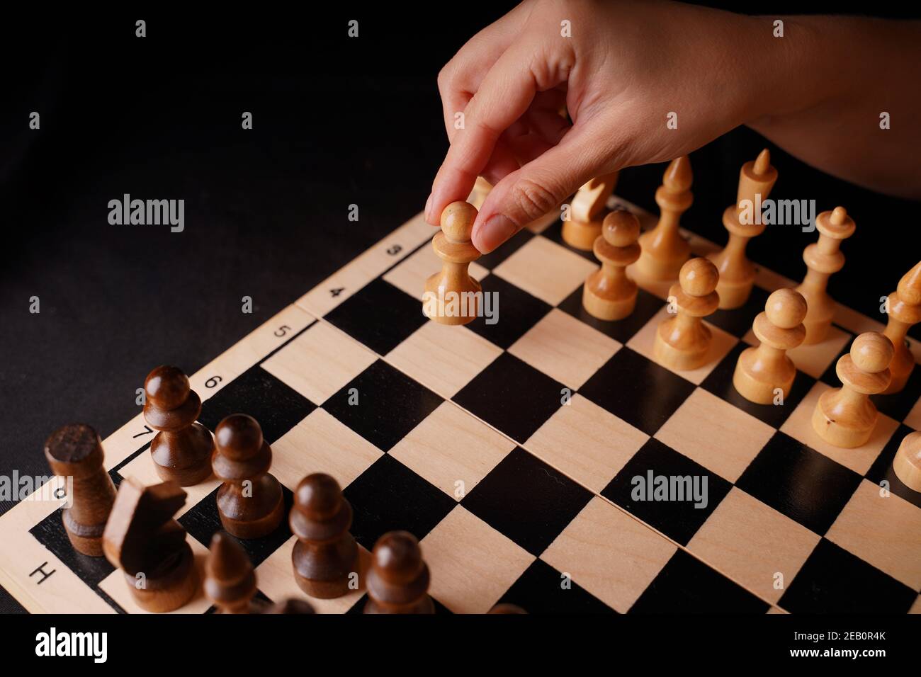 Close up of white and black wooden chess pieces on board. Woman's hand makes first move of white pawn on chessboard. Concept of intelligent, logical a Stock Photo