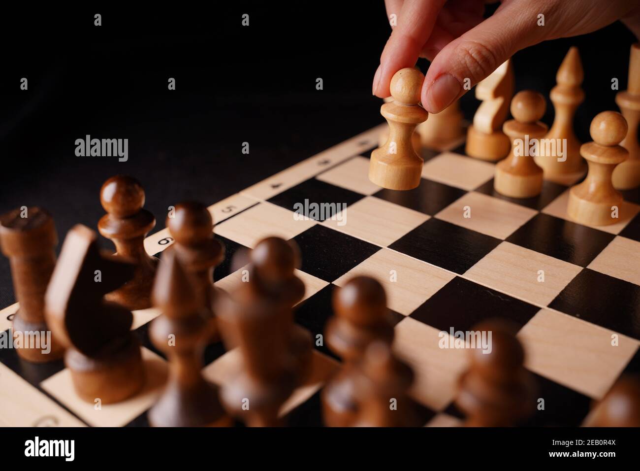 Close up of white and black wooden chess pieces on board. Woman's hand makes first move of white pawn on chessboard. Concept of intelligent, logical a Stock Photo