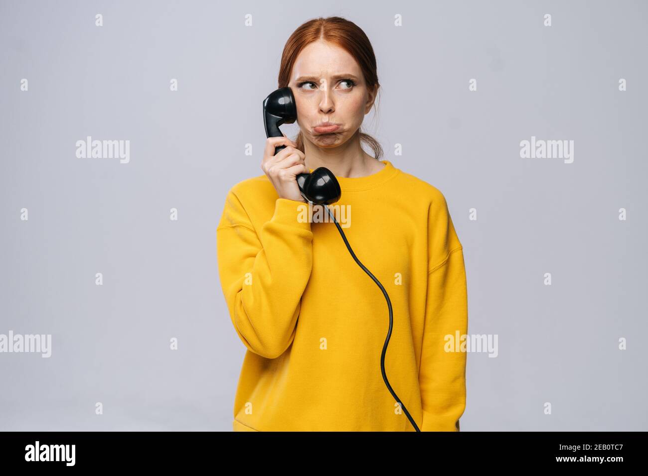 Confused young woman in stylish yellow sweater talking on retro phone and looking away Stock Photo