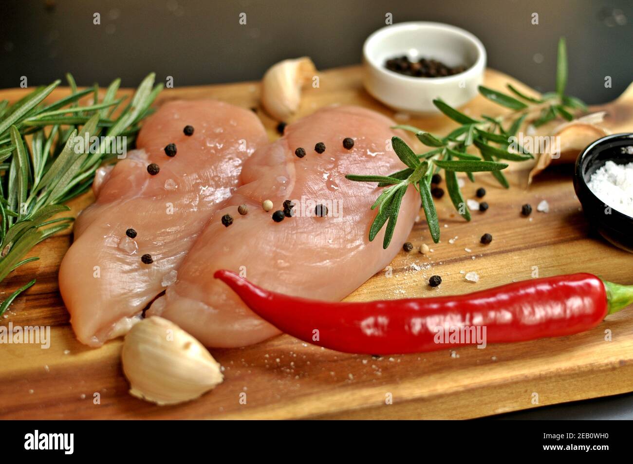 Raw fresh uncooked chicken breast meat fillet dish with rosemary, pepper, salt and red hot chili pepper on wooden board and black background. Stock Photo