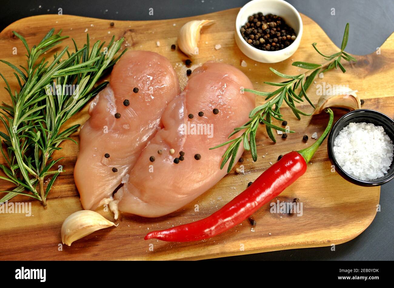 Raw fresh uncooked chicken breast meat fillet dish with rosemary, pepper, salt, red chilly pepper and garlic on wooden board and black background. Stock Photo