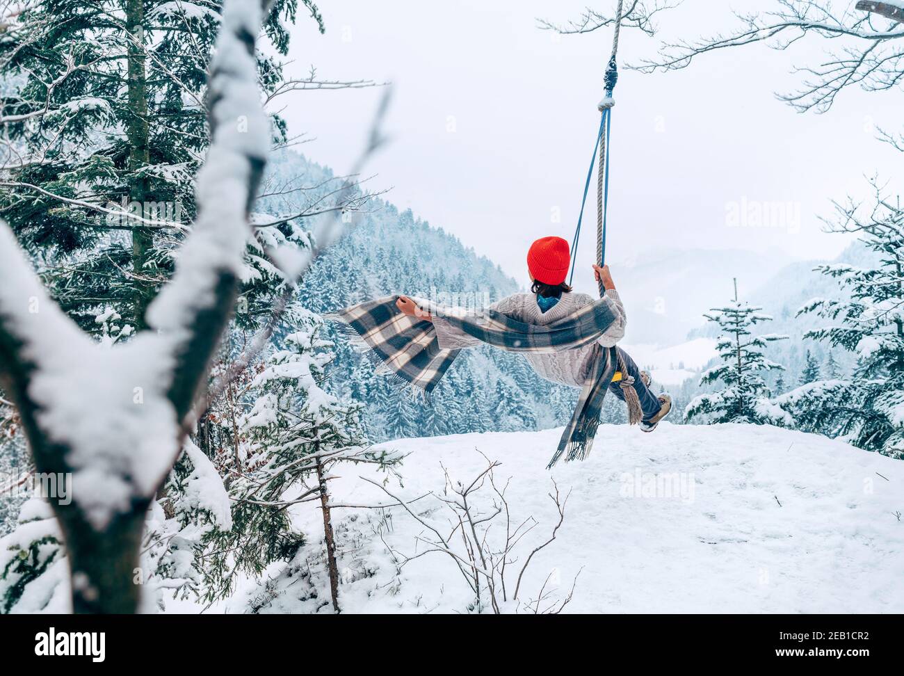 A young woman in warm clothes and Red Cap swinging on a swing between forest trees with picturesque snowy mountain view. Wintertime vacation concept i Stock Photo