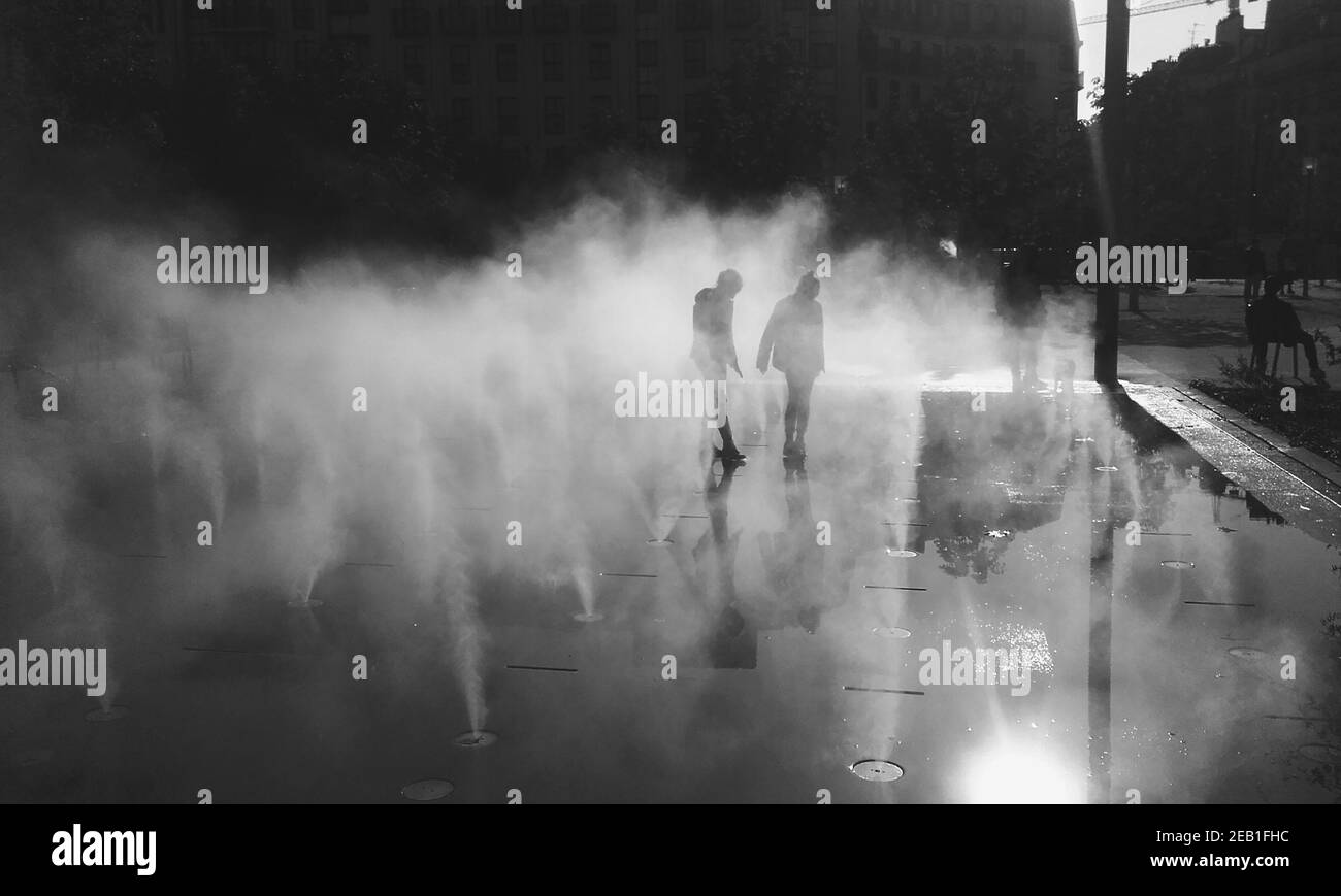 Blurry silhouettes of unrecognizable boy and girl playing in fountain water mist at autumn day. Les Halles square, Paris, France. Black and white phot Stock Photo