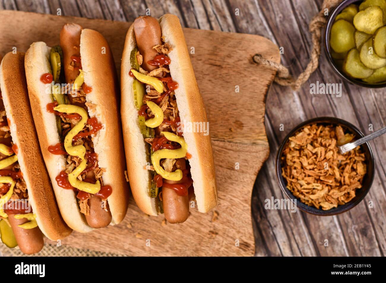 Homemade hotdogs with sausages and buns topped with preserved pickles, dried roast onions, mustard and ketchup next to bowls with ingredients Stock Photo