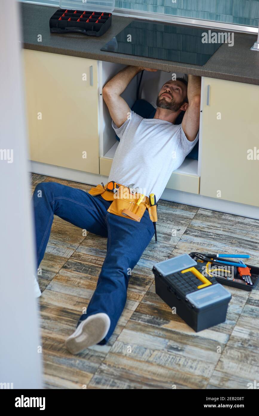 Stop leakage. Professional repairman, plumber wearing tool belt examining and fixing sink pipe in the kitchen indoors Stock Photo