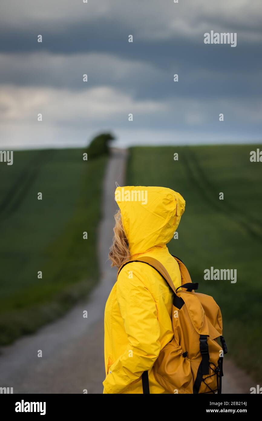 Rain is coming. Woman hiking on road and looking at cloudy sky. Backpacker wearing yellow waterproof jacket with hood Stock Photo