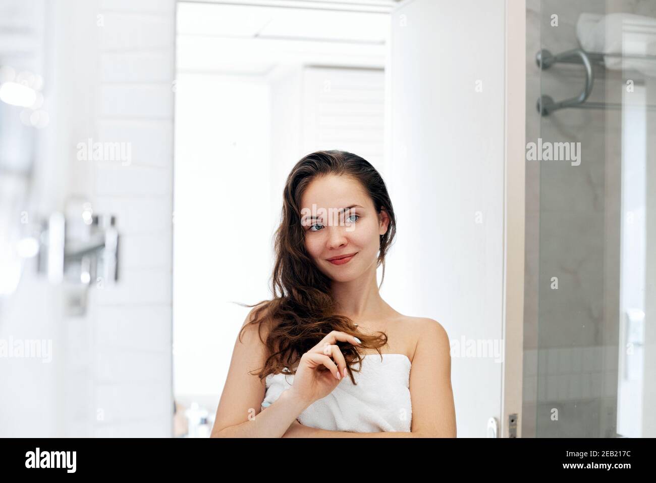 Hair and body care. Woman touching hair and smiling while looking in the mirror.Portrait of happy girl with wet hair in bathroom applying conditioner Stock Photo