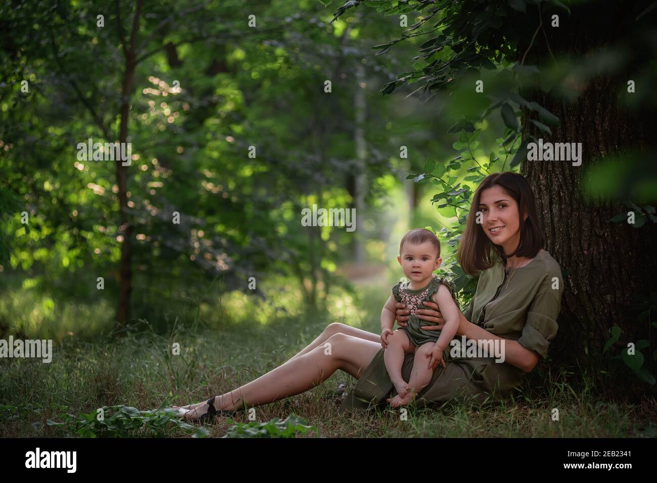 A healthy young mother holds a toddler baby in her arms. A happy family sits on the green grass, under a tall tree, plays, hugs, enjoys a walk in the Stock Photo