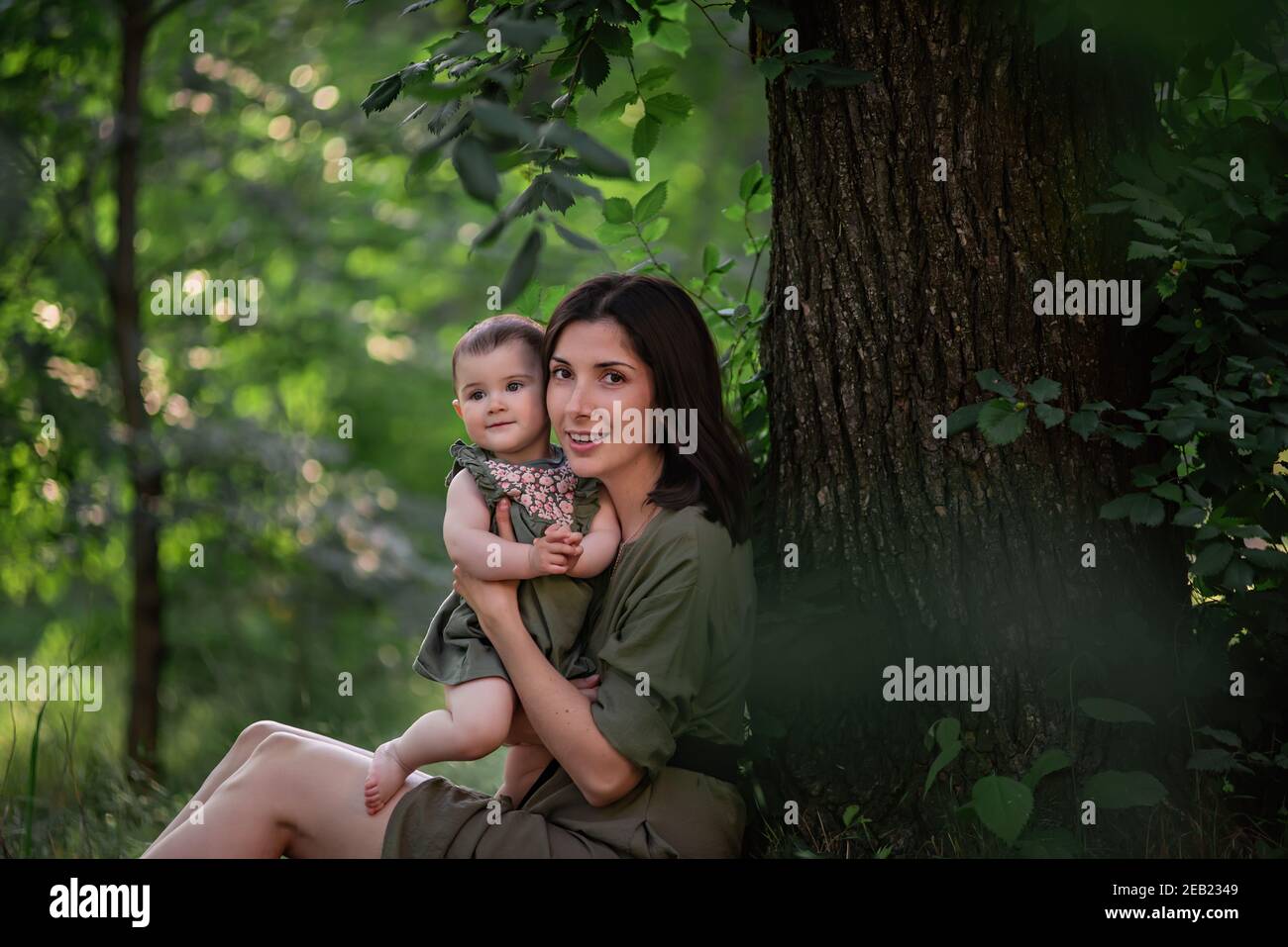 A healthy young mother holds a toddler baby in her arms. A happy family sits on the green grass, under a tall tree, plays, hugs, enjoys a walk in the Stock Photo