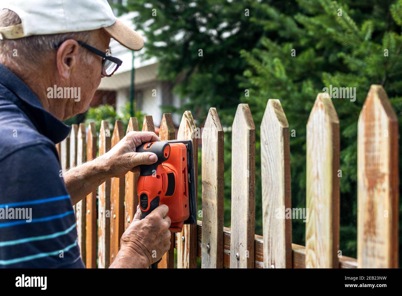 Sanding and preparing wooden fence for painting. Craftsperson restoring picket fence at backyard Stock Photo