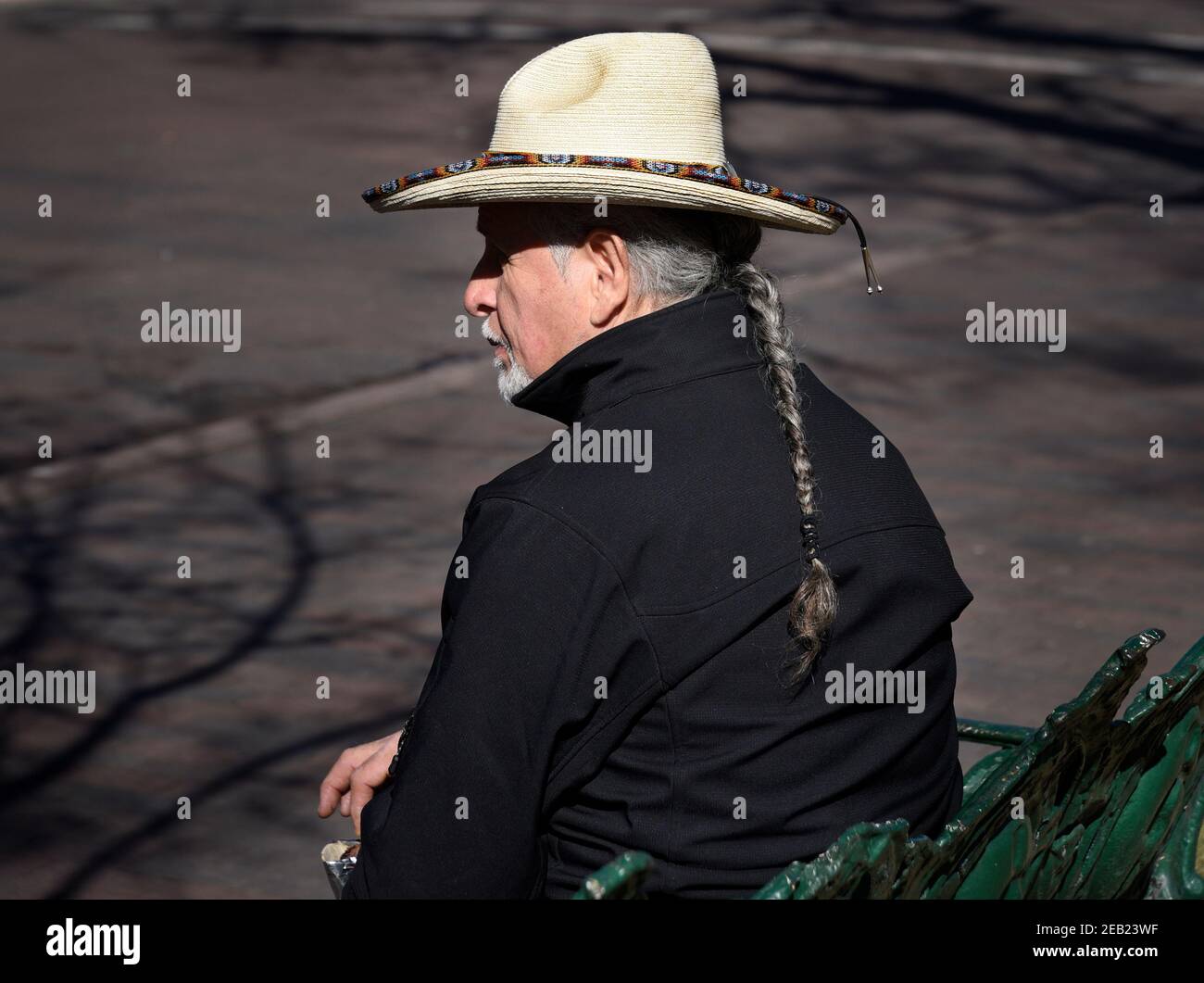 An Hispanic man with braided hair (Ramon Jose Lopez, a well-known artist) sits on a bench in the historic Plaza in Santa Fe, New Mexico. Stock Photo