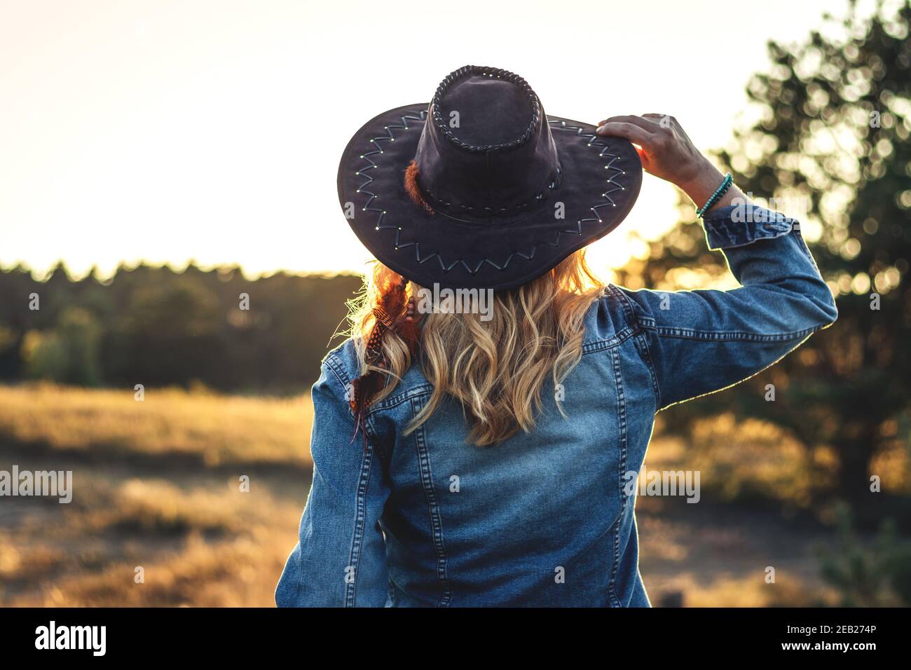 Blond hair woman with hat and denim jacket enjoying sunset outdoors. Fashion concept with woman wearing trendy casual clothing in nature at summer Stock Photo