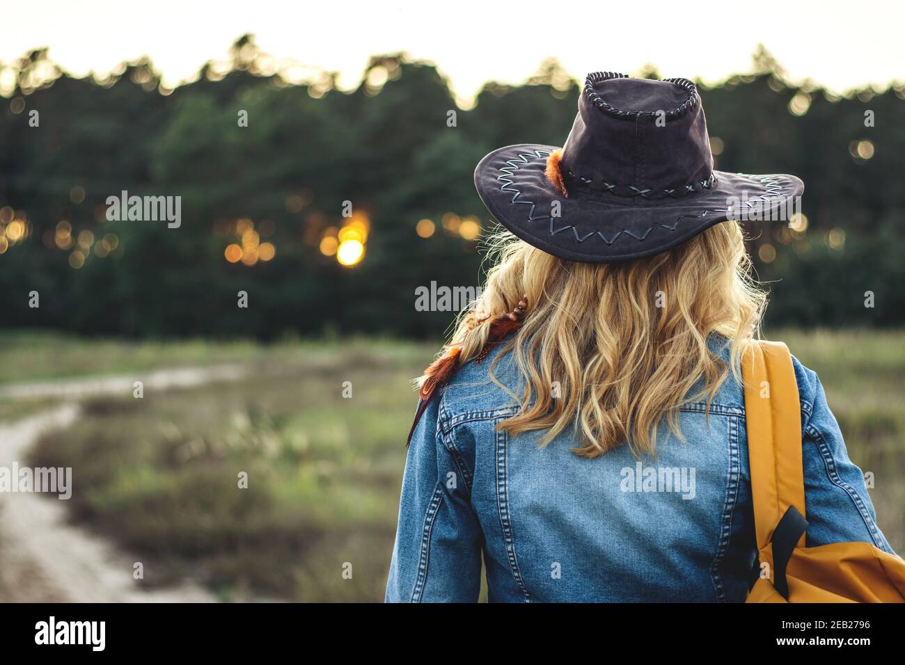 Woman tourist with hat and backpack hiking on footpath in nature during sunset. Rear view of blond hair female tourist wearing denim jacket is walking Stock Photo