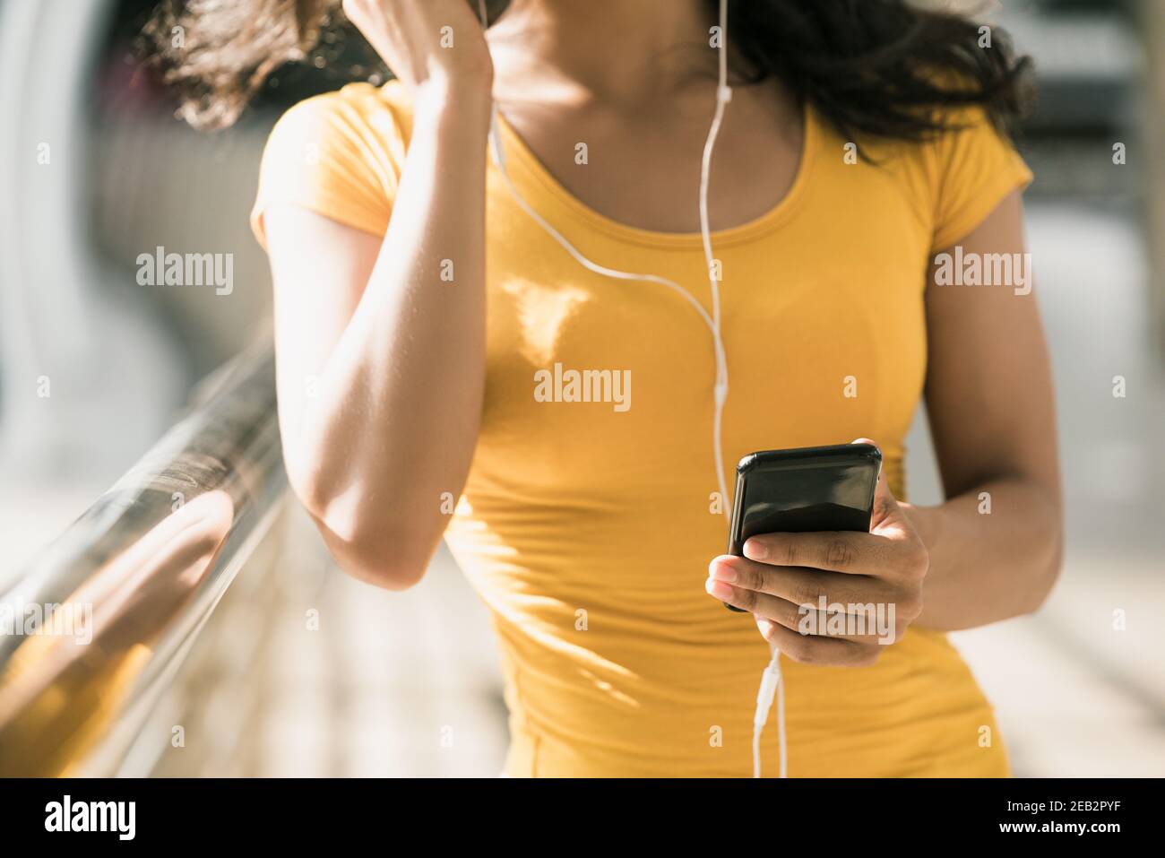 Young woman wearing earphones listening to music from smartphone while walking in the city Stock Photo
