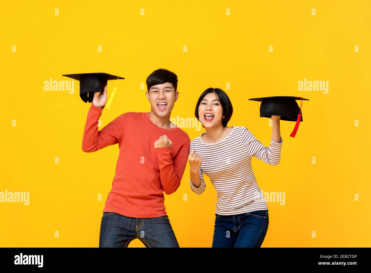 Excited happy Asian college students holding graduate caps in colorful yellow studio background Stock Photo