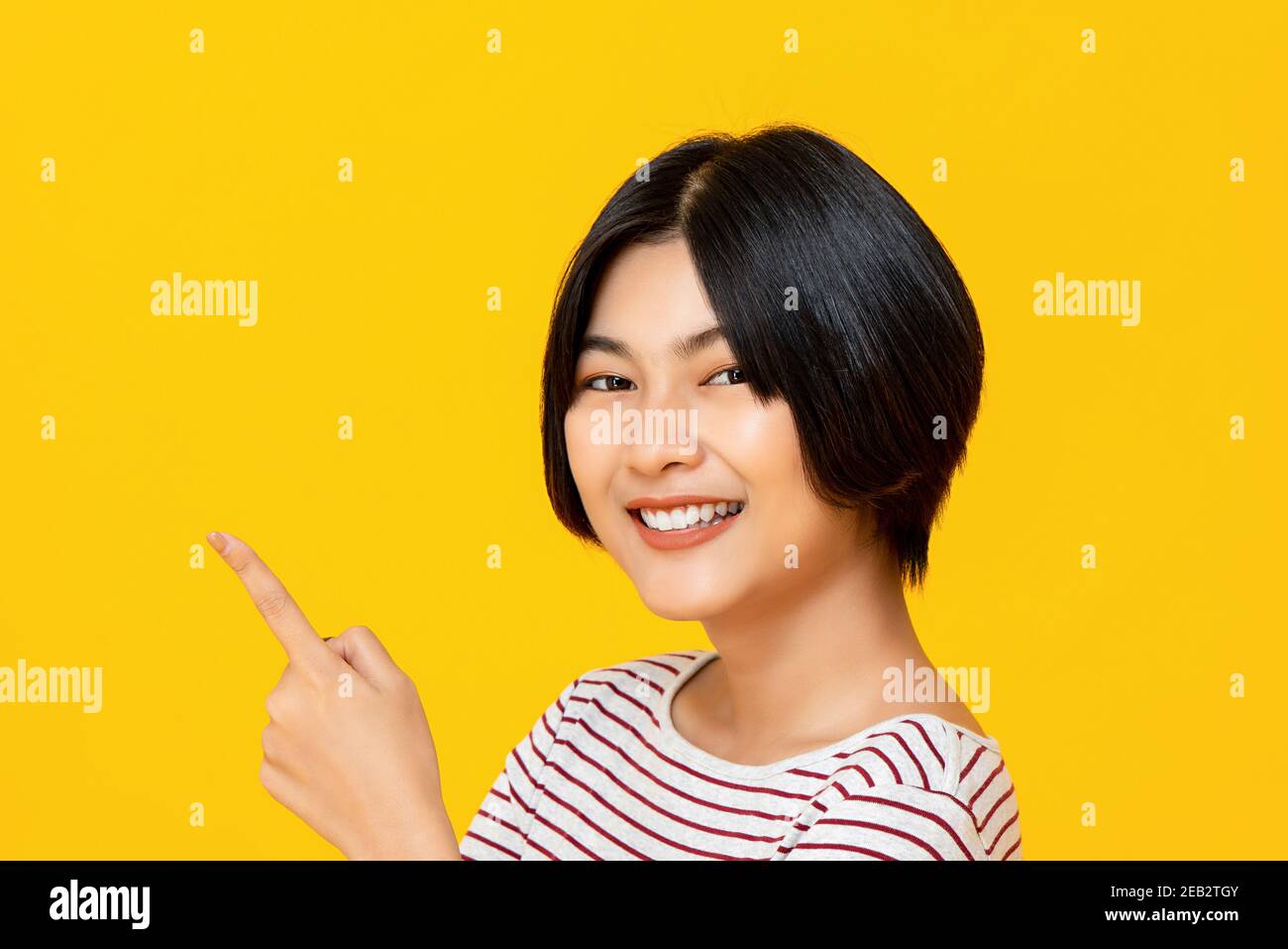 Beautiful smiling young Asian woman with short hairstyle pointing hand to empty space on yellow background Stock Photo