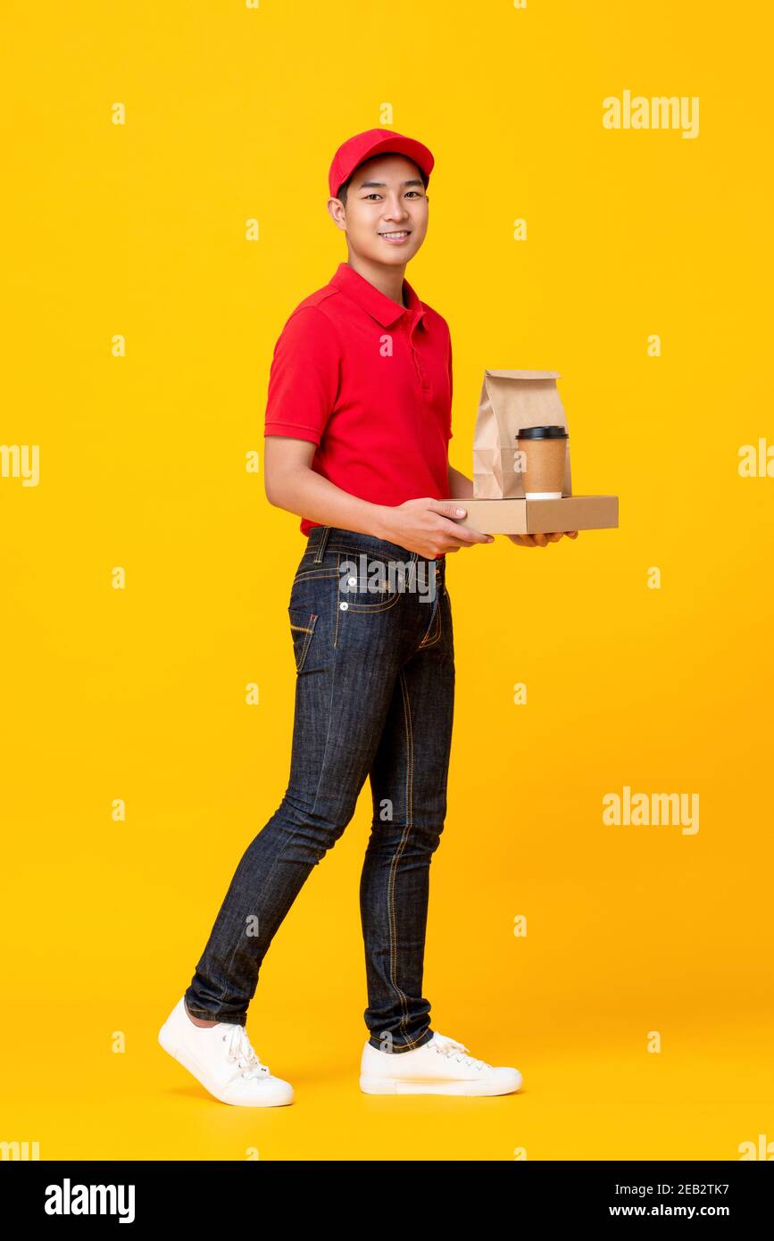 Young Asian male fast food crew in red uniform delivering take away meal in yellow isolated studio background Stock Photo