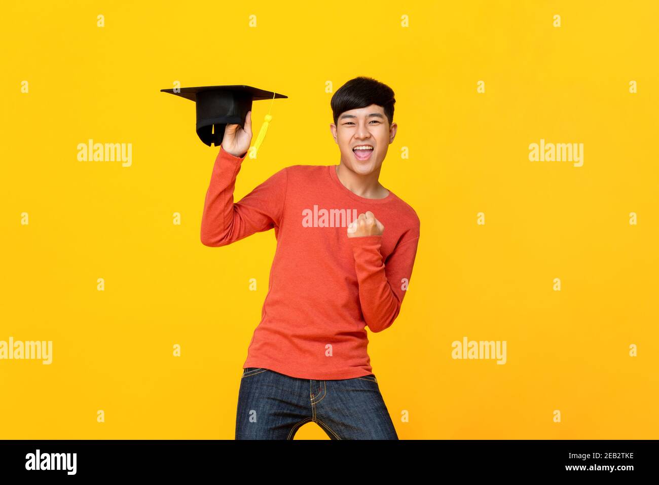 Excited young asian male student in casual attire holding a graduation cap doing winning closed fist gesture in yellow isolated studio background Stock Photo