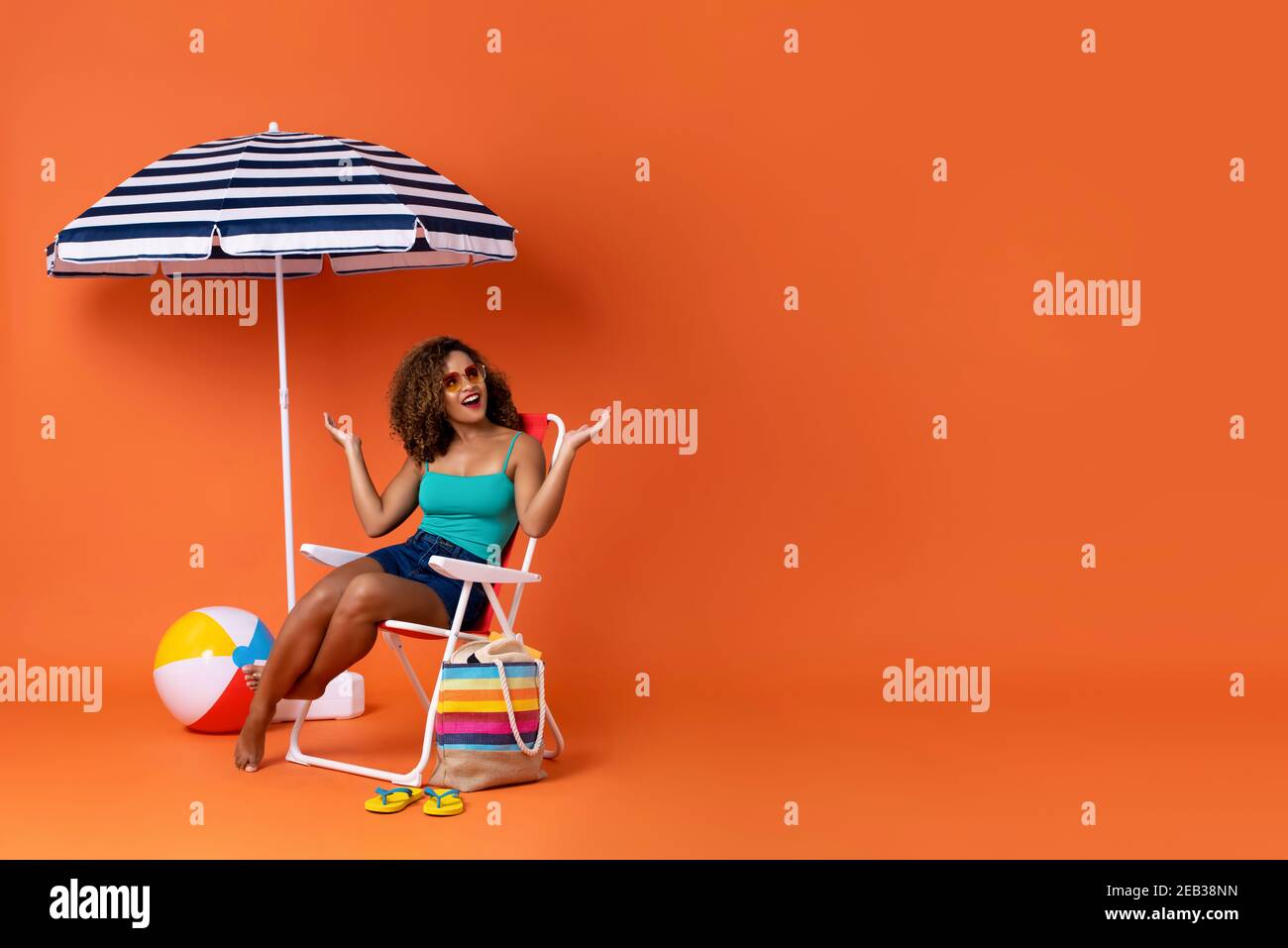Beautiful African American woman sitting on a beach chair with colorful accessories and  umbrella in studio summer orange background with copy space Stock Photo