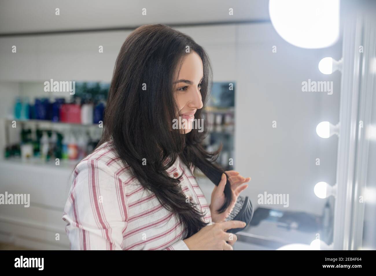 Long-haired young brunette after hair styling looking in the mirror and feeling contented Stock Photo