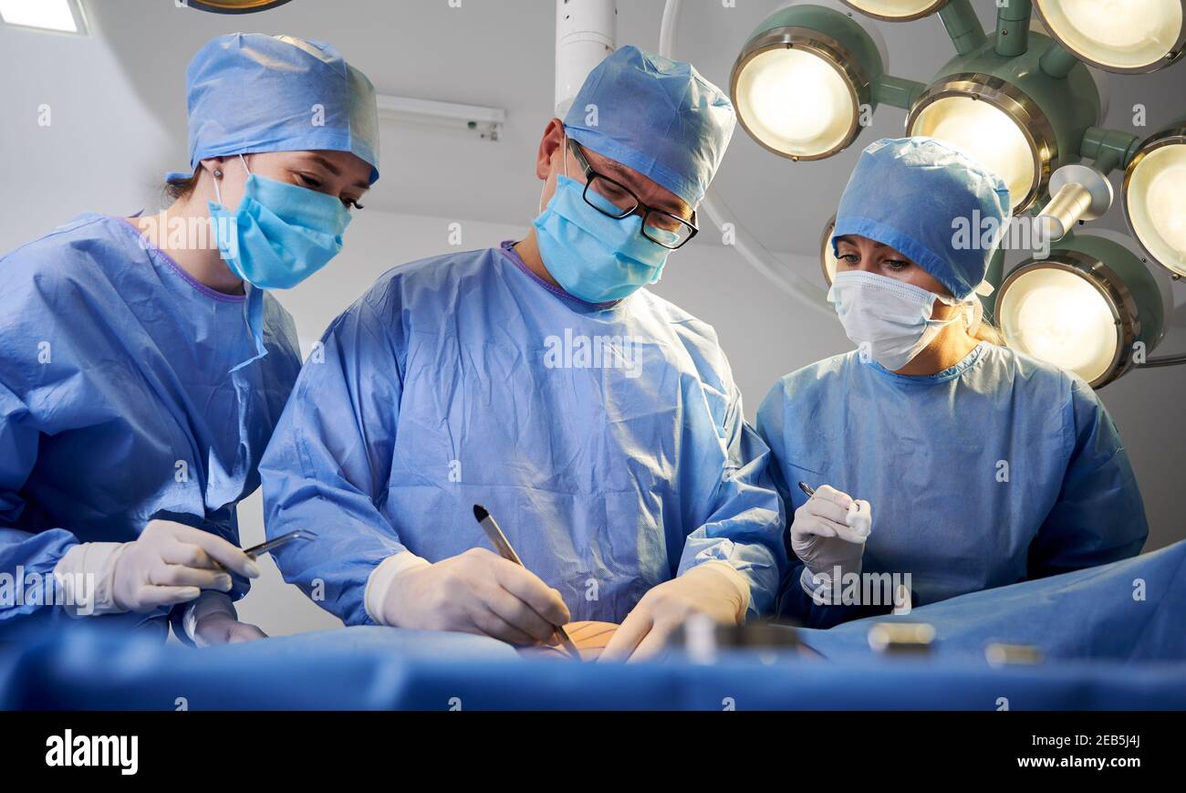 Doctors performing aesthetic surgery while patient lying on operating table. Surgeon and assistants performing cosmetic surgery in operating room. Concept of medicine, abdominoplasty, plastic surgery. Stock Photo
