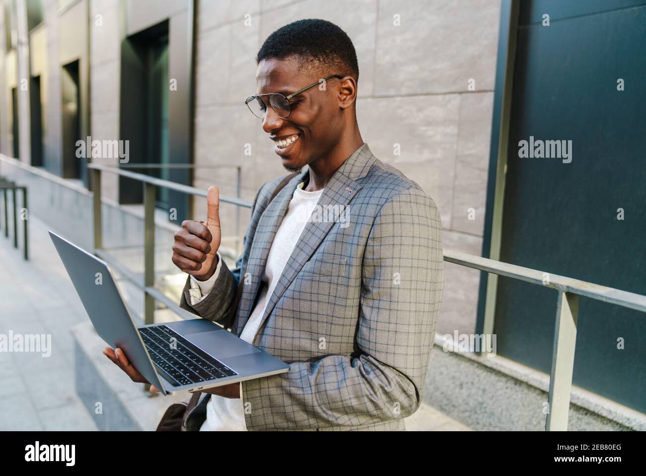 Joyful african american man showing thumb up and using laptop while standing outdoors Stock Photo