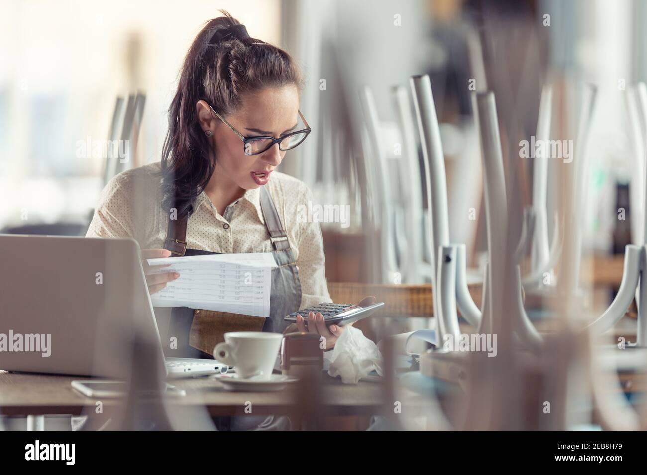 Unhappy cafe owner does tax calculations and paperwork over coffee and open laptop. Stock Photo