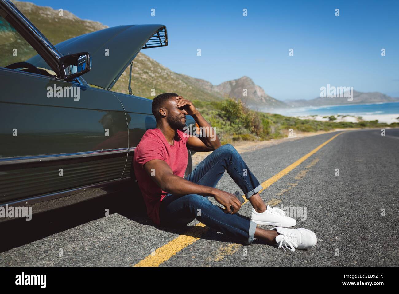 African american man holding smartphone sitting on road beside broken-down car with open bonnet Stock Photo