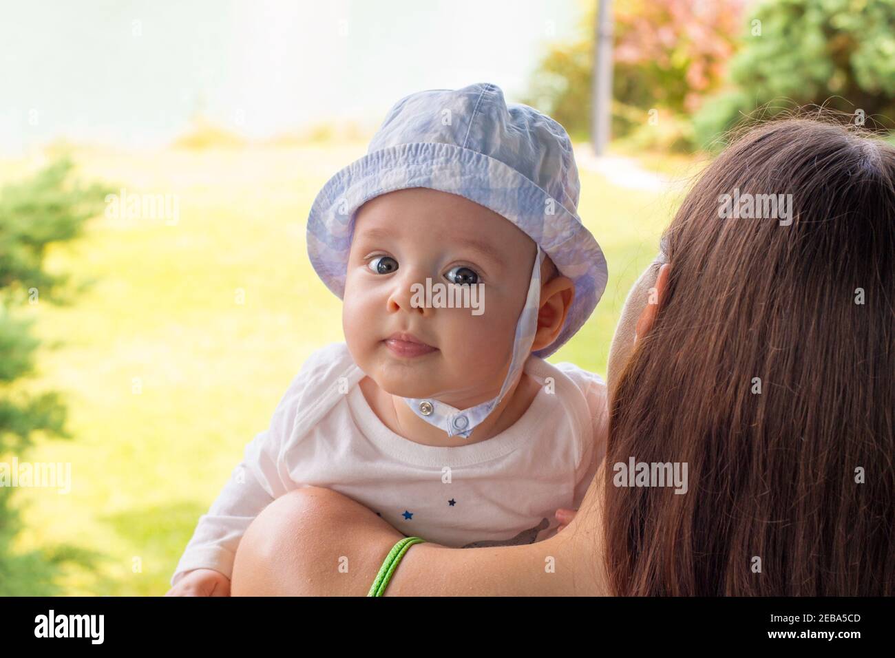 Blue eyed baby in hat resting on pretty mother’s shoulder, infant face portrait close up Stock Photo