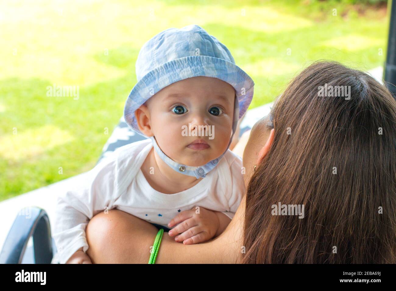 New mother and child on deckchair at backyard with green grass garden view and holding her little baby, baby boy resting on mother’s shoulder and look Stock Photo