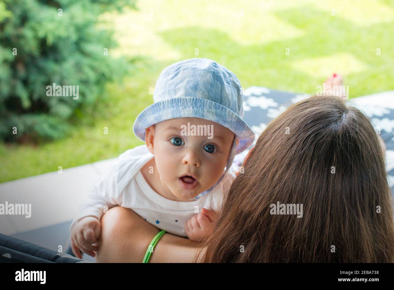 Surprised baby face with fascinated expression, little cute child in mother's arms looks out with open mouth and focused wide open blue eyes Stock Photo