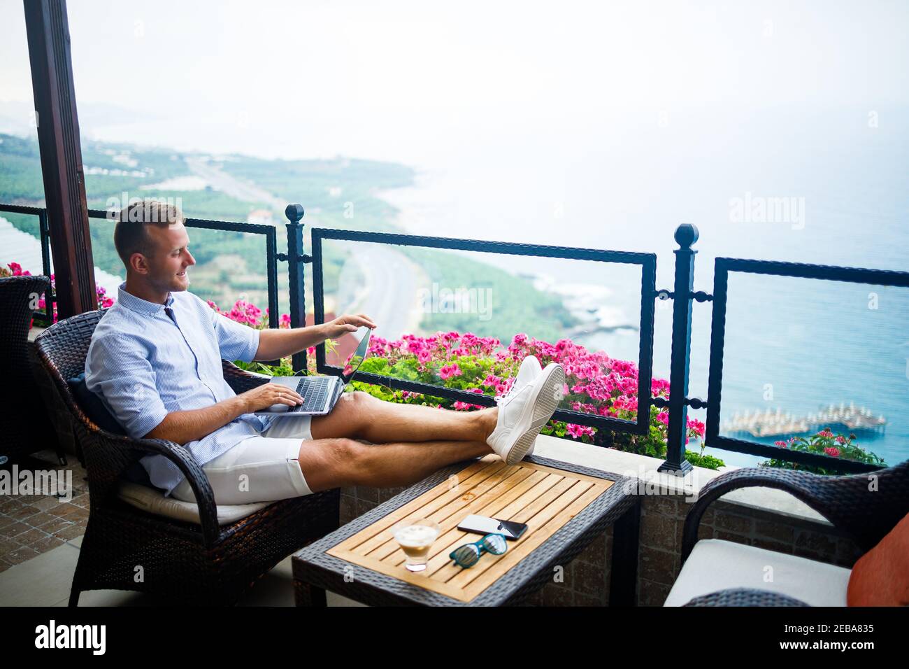 Young handsome businessman in a white shirt and shorts sits with a laptop in a cafe at the table. Work while resting Stock Photo