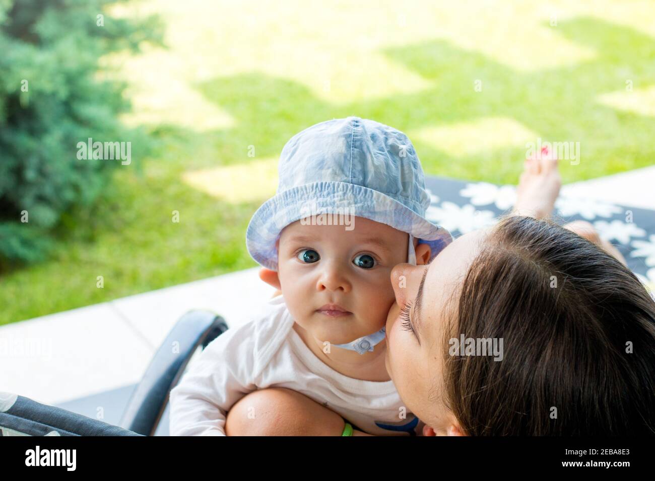 Cheerful mother hugging, cuddling and cheek kissing child outdoor, portrait of lovely blue eyed baby boy resting in mom's loving arms Stock Photo