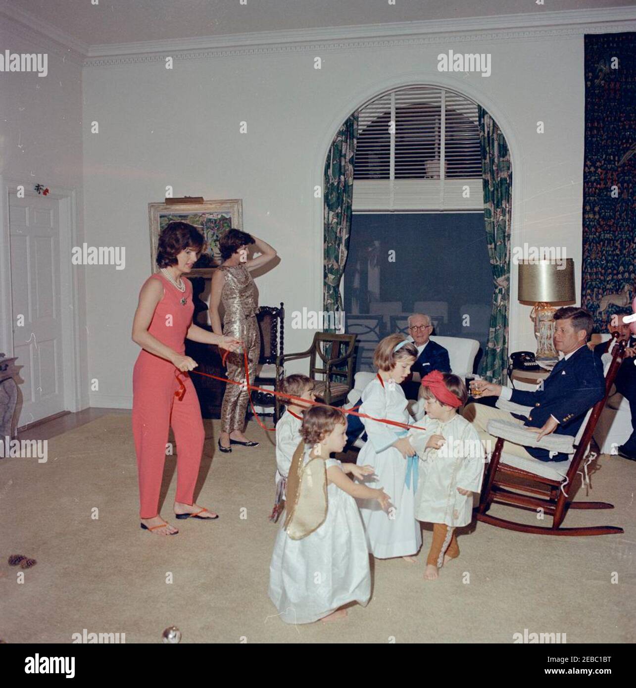 Christmas Day, Palm Beach. President John F. Kennedy celebrates Christmas with his family at the residence of C. Michael Paul in Palm Beach, Florida. Left to right: First Lady Jacqueline Kennedy (holding red ribbons); Eunice Kennedy Shriver; John F. Kennedy, Jr.; Anna Christina Radziwill (in foreground); Caroline Kennedy; Joseph P. Kennedy, Sr. (seated in armchair); Anthony Radziwill; President Kennedy (seated in rocking chair); Prince Stanislaus Radziwill of Poland (at edge of frame). [Notes: Blemishes throughout image are original to the negative.] Stock Photo