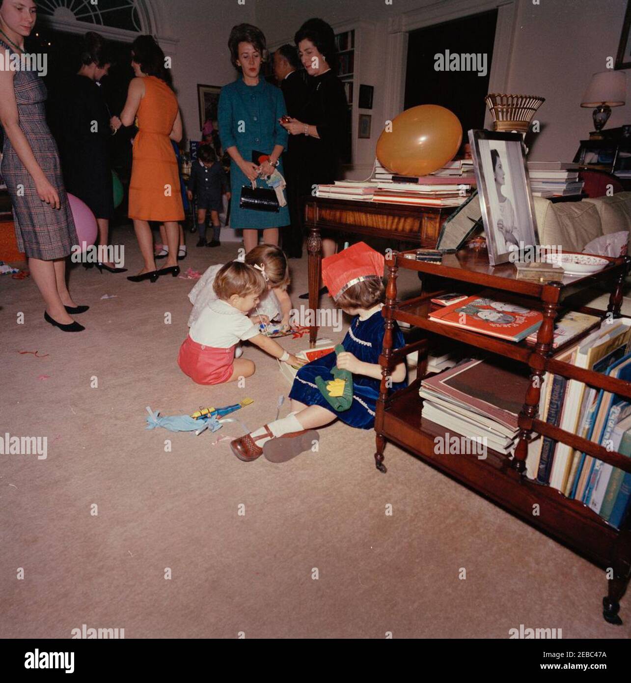 Birthday party for Caroline Kennedy and John F. Kennedy, Jr.. Caroline Kennedy and John F. Kennedy, Jr. (both seated at center left), play in the West Sitting Hall of the White House, Washington, D.C., during a joint birthday party in their honor; First Lady Jacqueline Kennedy (back to camera) stands at left in background. Also pictured: Janet Auchincloss; White House butler, John W. Ficklin. Stock Photo