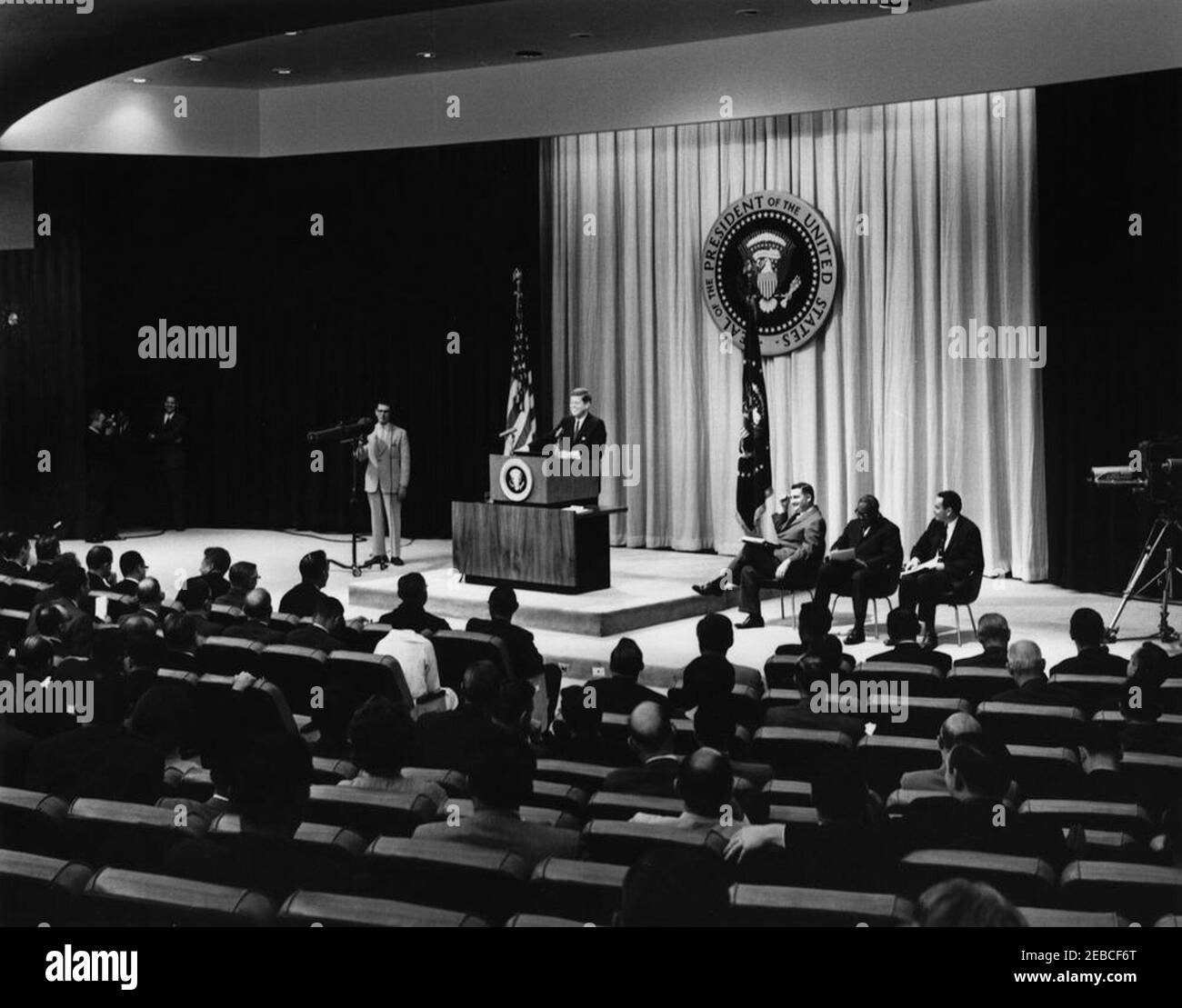 Press Conference, State Department Auditorium, 4:00PM. President John F. Kennedy laughs while standing at lectern during Press Conference. Seated onstage at right (L-R): Press Secretary Pierre Salinger; Associate Press Secretary Andrew Hatcher; Deputy Press Secretary Malcolm Kilduff, Jr. State Department Auditorium, Washington, D.C. Stock Photo