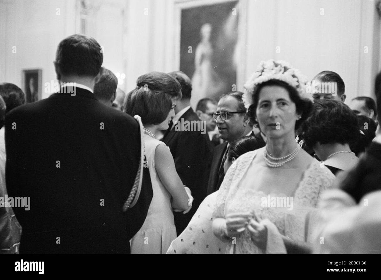 Diplomatic Reception, 6:00PM. First Lady Jacqueline Kennedy (center) visits with Ambassador of Nepal, Matrika Prasad Koirala, in the East Room during a diplomatic reception at the White House, Washington, D.C. President John F. Kennedy (mostly hidden, back to camera) stands behind Mrs. Kennedy. Naval Aide to the President, Captain Tazewell Shepard, stands at left in foreground (back to camera); Military Aide to the President, General Chester V. Clifton, stands at right (face obscured). Stock Photo