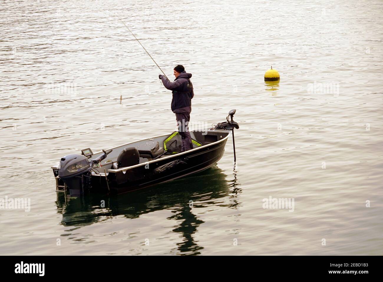 A hobby fisherman attaching bait on the hook on the lake Zurich in winter. He is standing in a boat and there is a yellow buoy on the water surface. Stock Photo