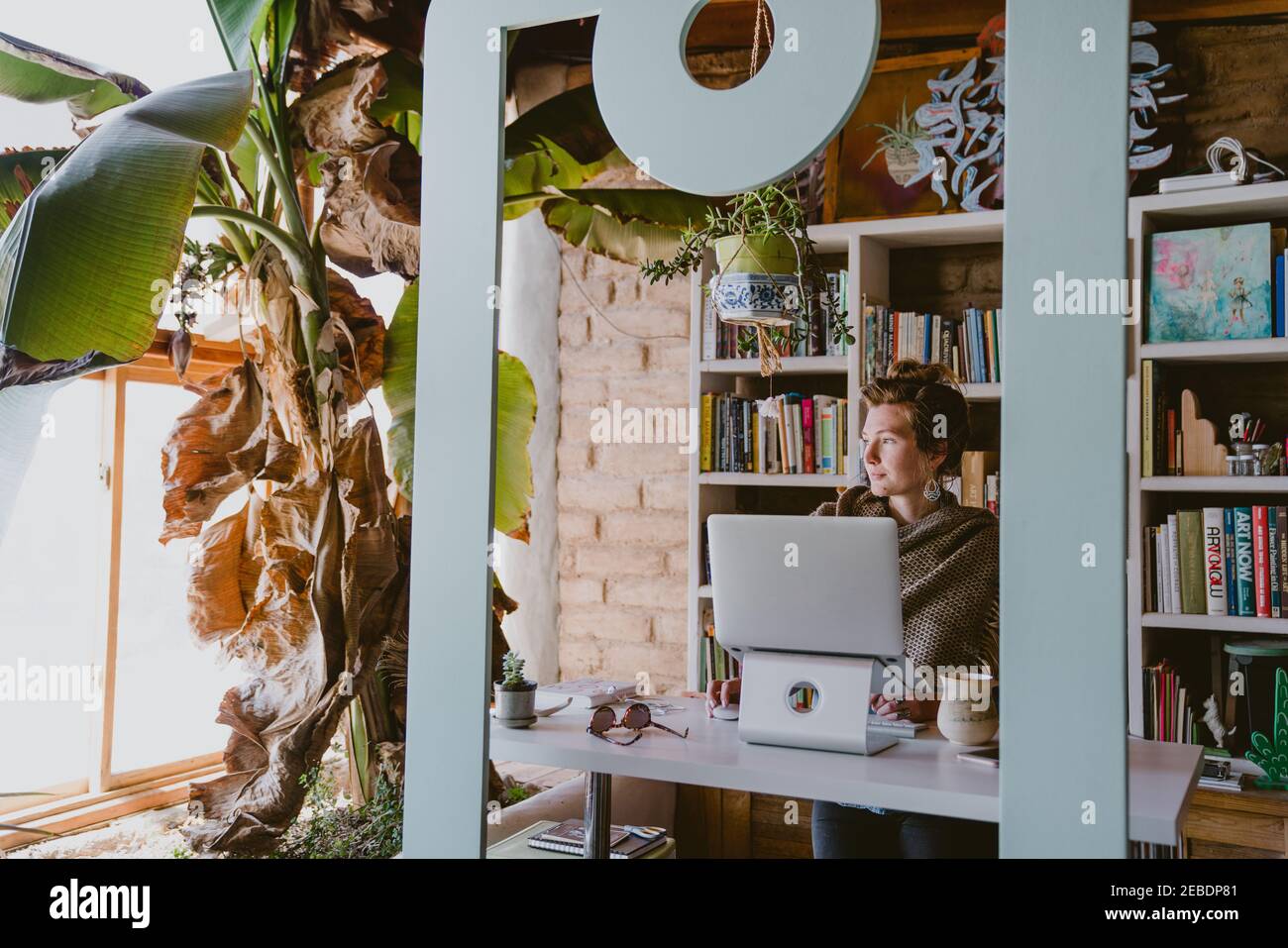 Woman works on laptop from home office surrounded by books and plants Stock Photo