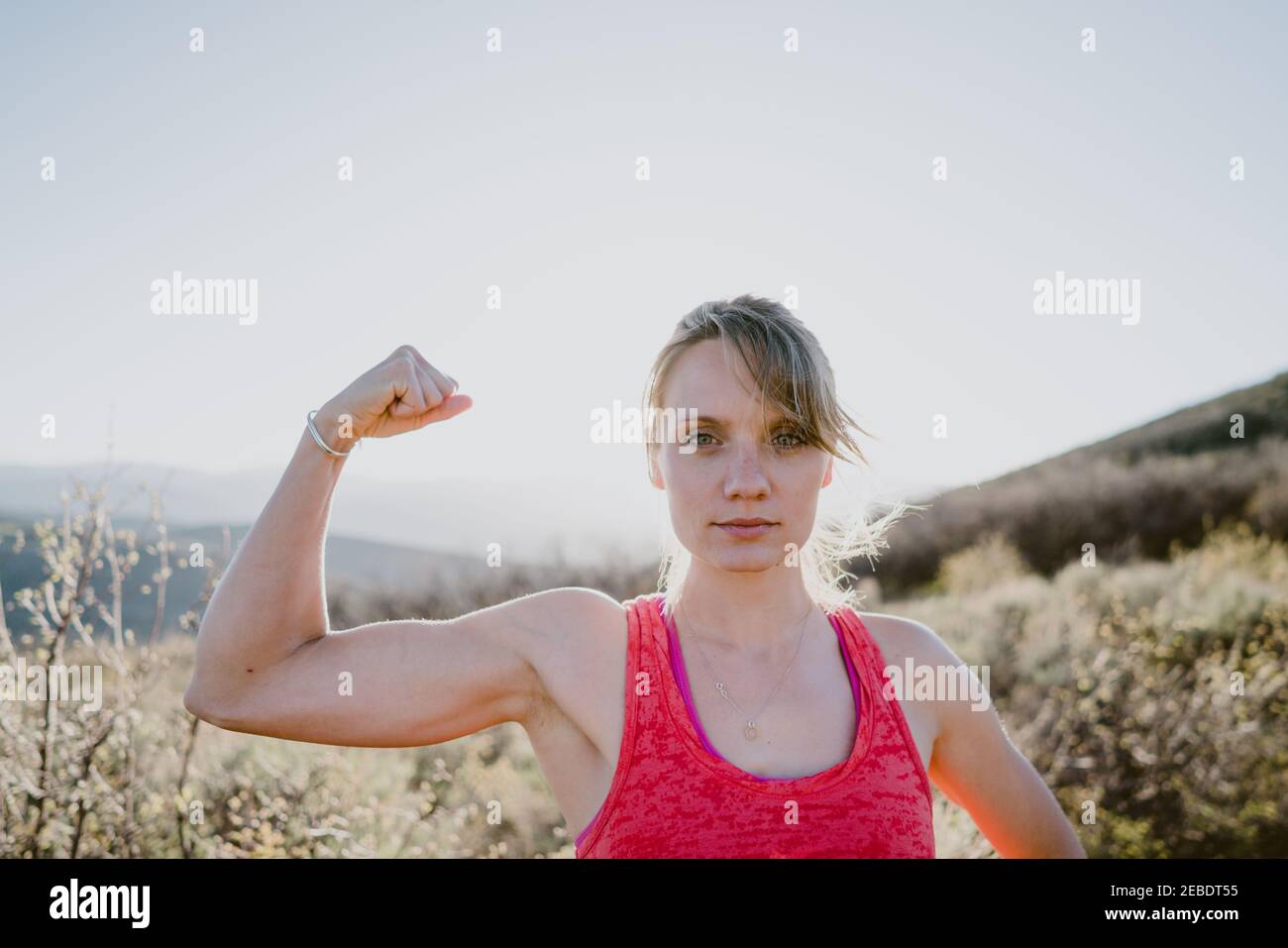 Athletic blonde woman flexes muscles with sun and mountains behind Stock Photo