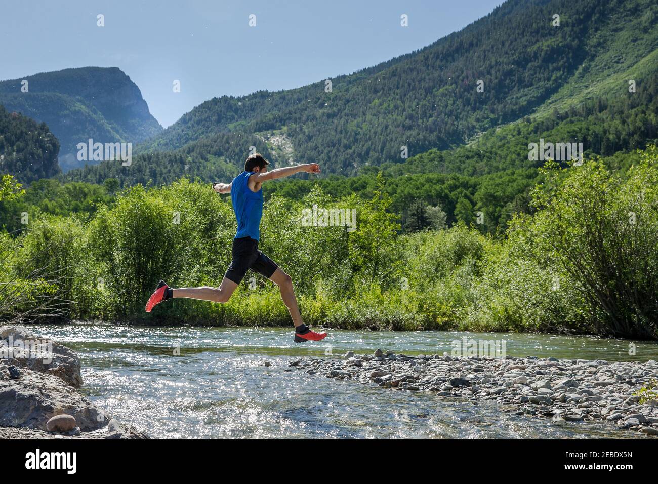 Male trail runner leaps over rocky river on sunny day in the mountains Stock Photo