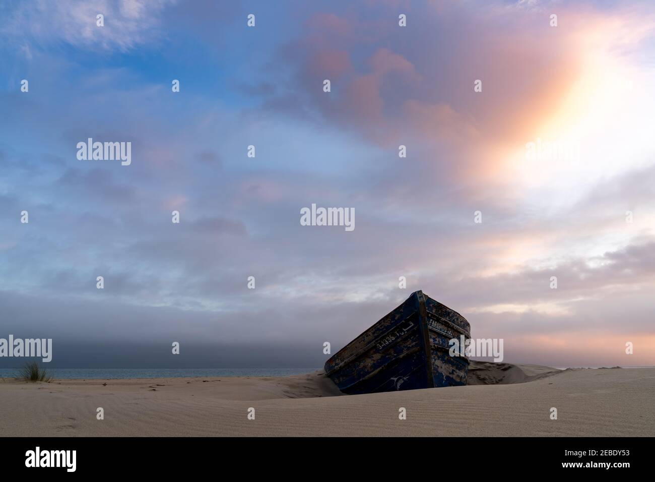 colorful wreck of an old wooden rowboat buried in the sand with a beautiful sunset sky and ocean behind Stock Photo