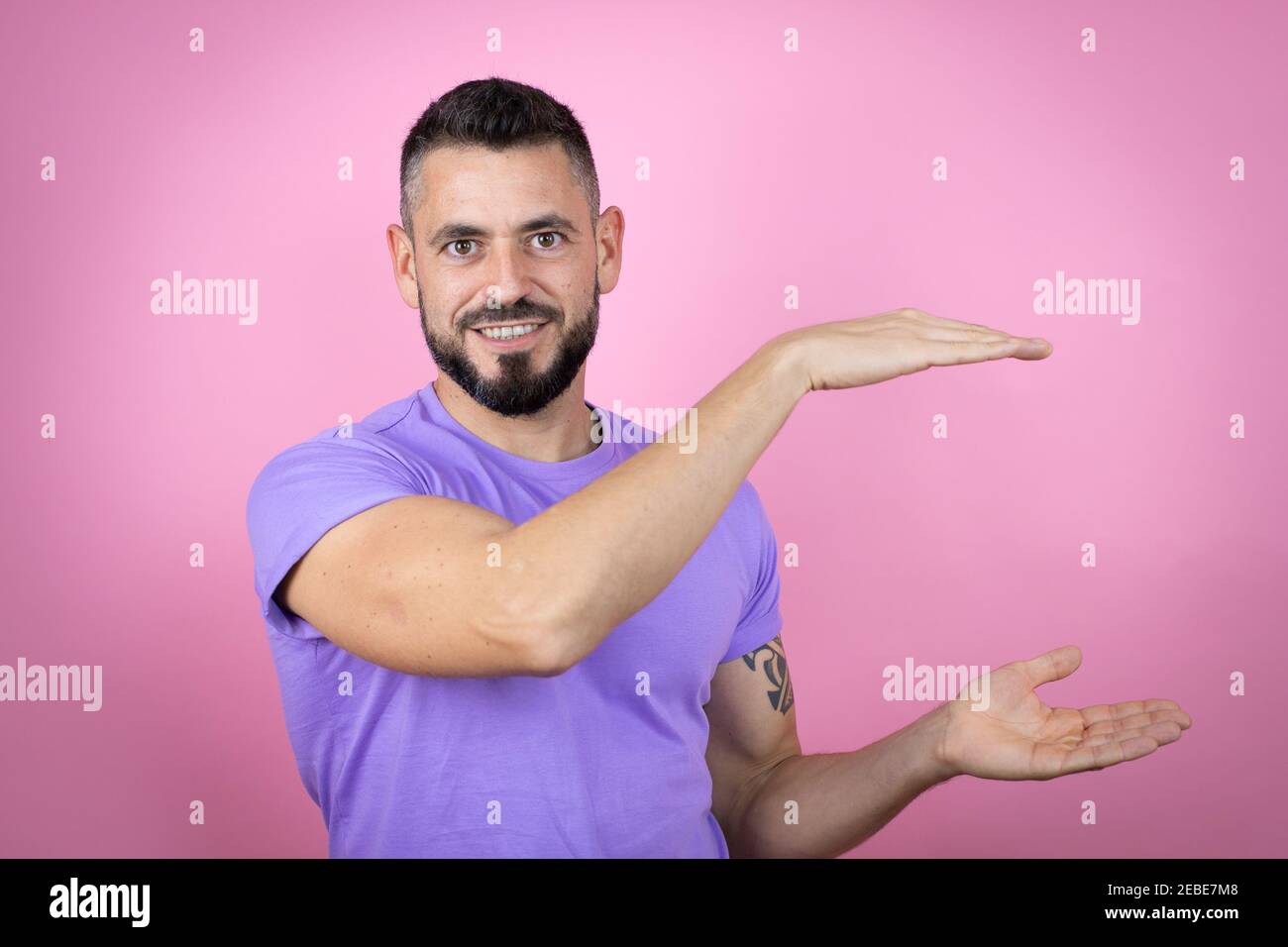 Young handsome man wearing casual t-shirt over pink background gesturing with hands showing big and large size sign, measure symbol. Smiling . Measuri Stock Photo