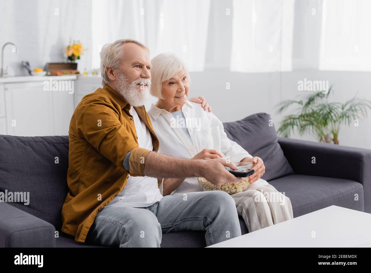 Smiling elderly couple watching tv with popcorn on couch Stock Photo