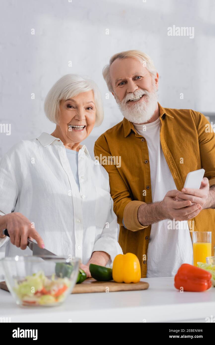 Smiling senior couple with smartphone looking at camera while cooking on blurred foreground Stock Photo