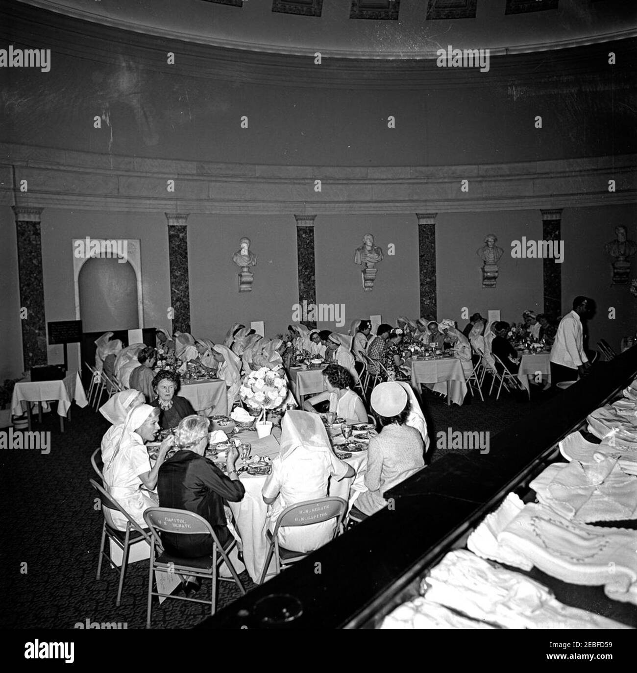 First Lady Jacqueline Kennedy (JBK) attends the Senate Ladies Red Cross Unit Luncheon. First Lady Jacqueline Kennedy (back to camera, wearing hat), Lady Bird Johnson, and members of the Senate Ladies Red Cross Unit attend a luncheon in the Old Supreme Court Chamber, United States Capitol Building, Washington, D.C. The Red Cross unit (also known as u201cLadies of the Senateu201d) is comprised of the wives of members of the US Senate. Stock Photo