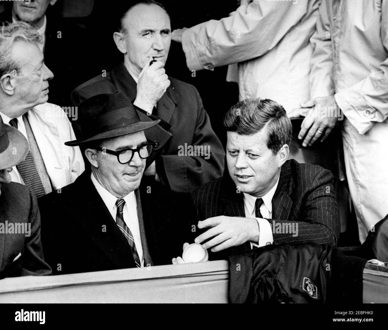 Opening Day of the 1961 Baseball Season, 1:10PM. President John F. Kennedy and Special Assistant to the President Dave Powers look at a baseball during the opening game of the 1961 baseball season at Griffith Stadium, Washington, D.C. Also pictured are Senate Minority Leader Everett Dirksen of Illinois (behind Powers) and Senate Majority Leader Mike Mansfield of Montana (behind President Kennedy). Stock Photo