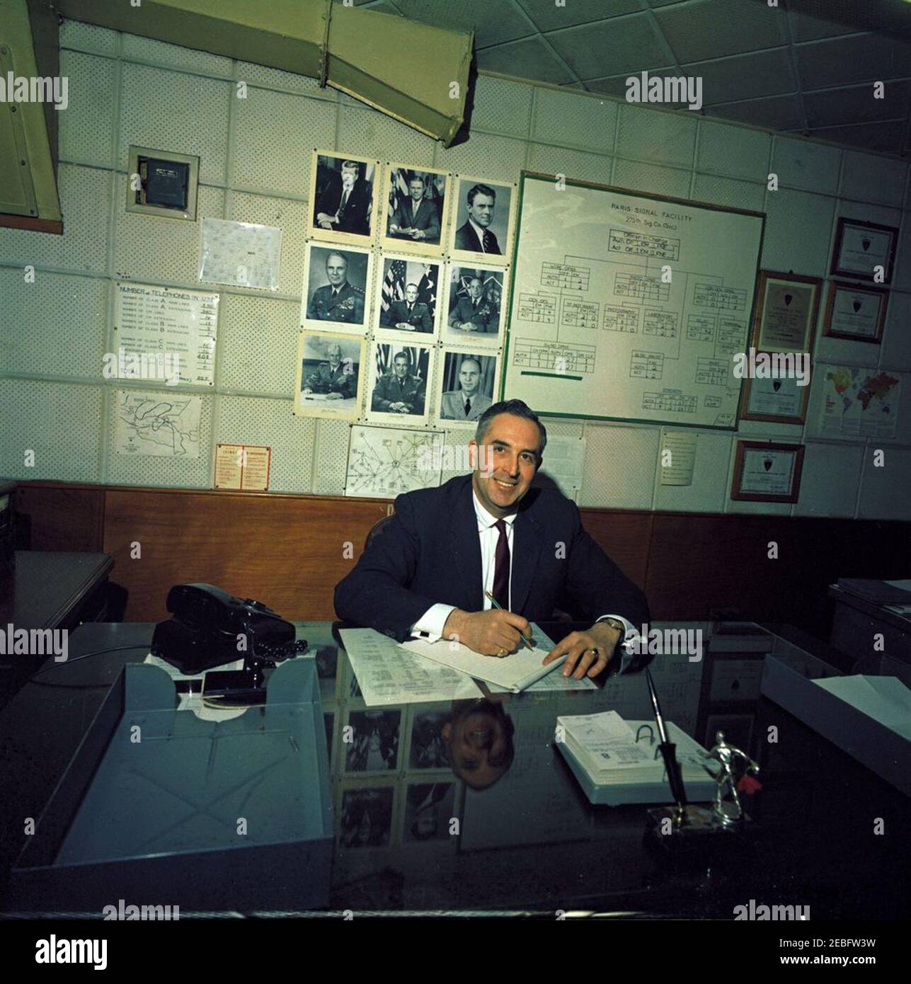 White House Army Signal Agency (WHASA) advance trip to Paris, France. White House Army Signal Agency (WHASA) advance trip to Paris. Unidentified man sitting at desk. Poster on wall reads: u201cParis Signal Facility, 275th Sig. Co. (Svc.),u201d with a grid of staff assignments listed below. Unidentified building, Paris, France. Stock Photo
